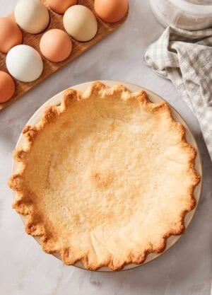 Overhead shot of a blind-baked crust in a pie dish with eggs and a checkered linen off to the side.
