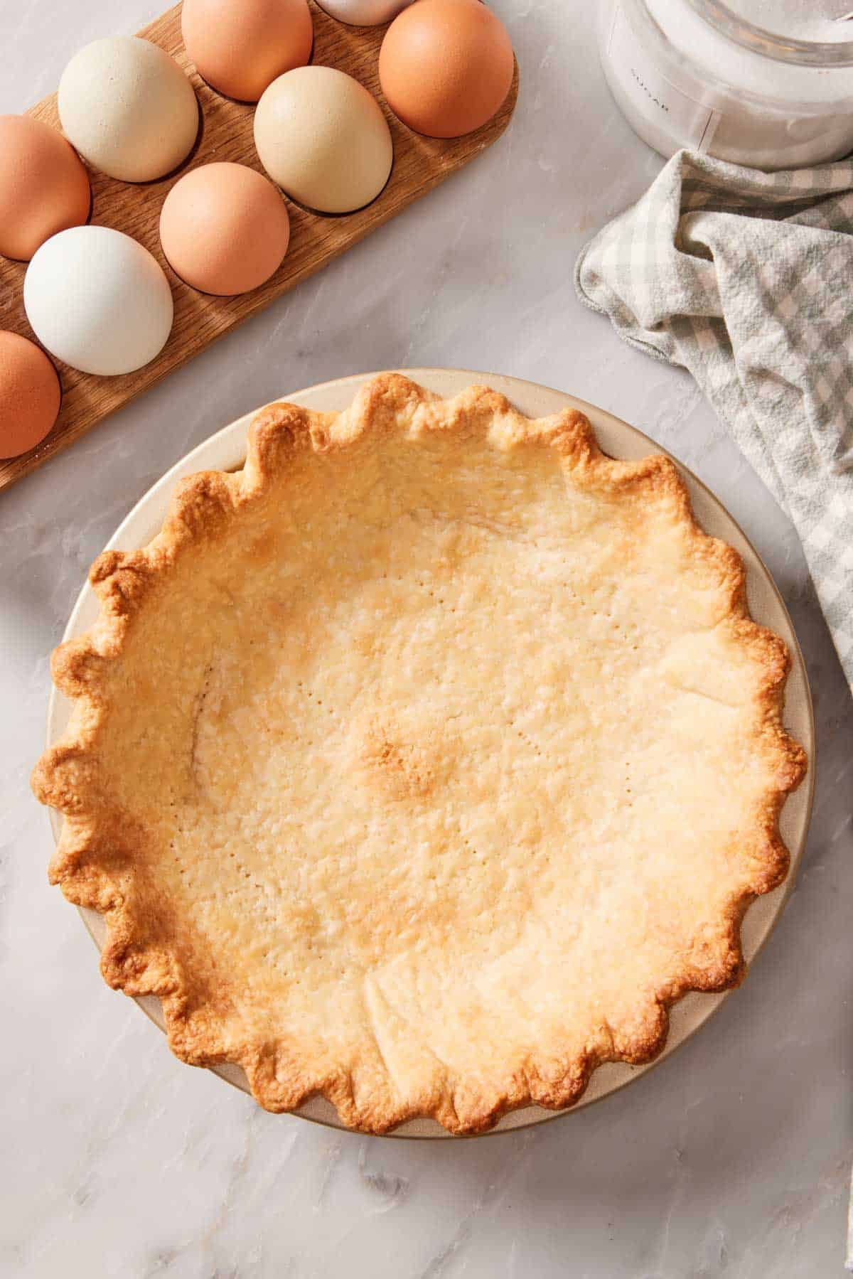 Overhead shot of a blind-baked crust in a pie dish with eggs and a checkered linen off to the side.