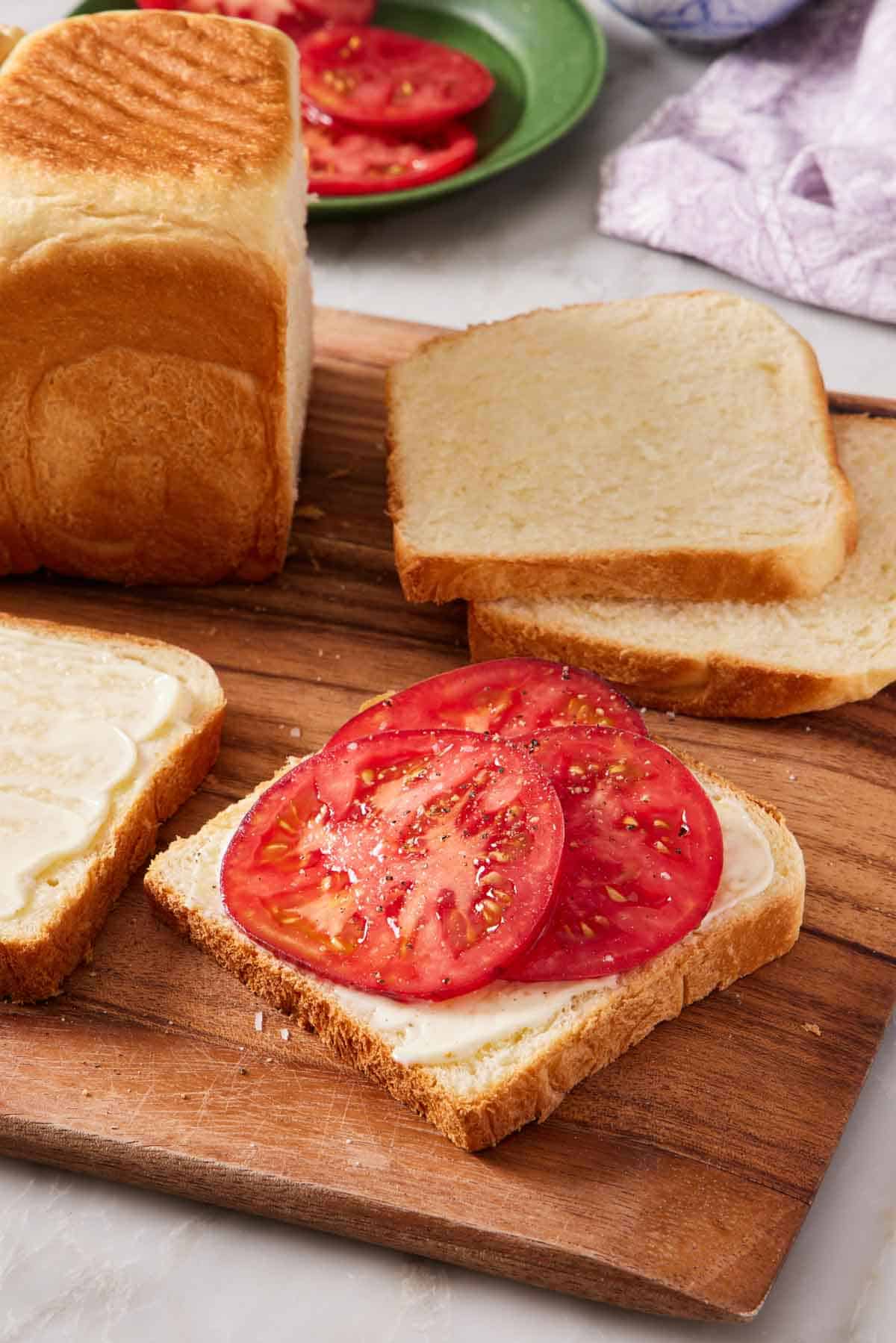 Slices of milk bread on a wooden cutting board, one with sliced tomato on it.