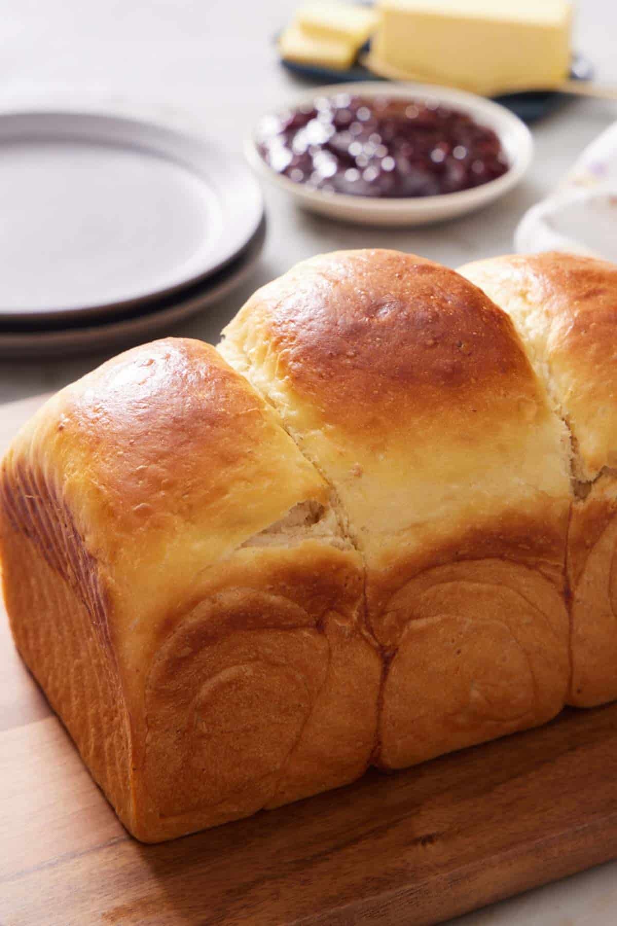 A loaf of milk bread mostly in frame on a wooden cutting board, with butter and jam out of focus in the background.