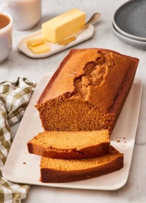 A partially sliced loaf of pumpkin bread on a platter with a stick of butter in the background.