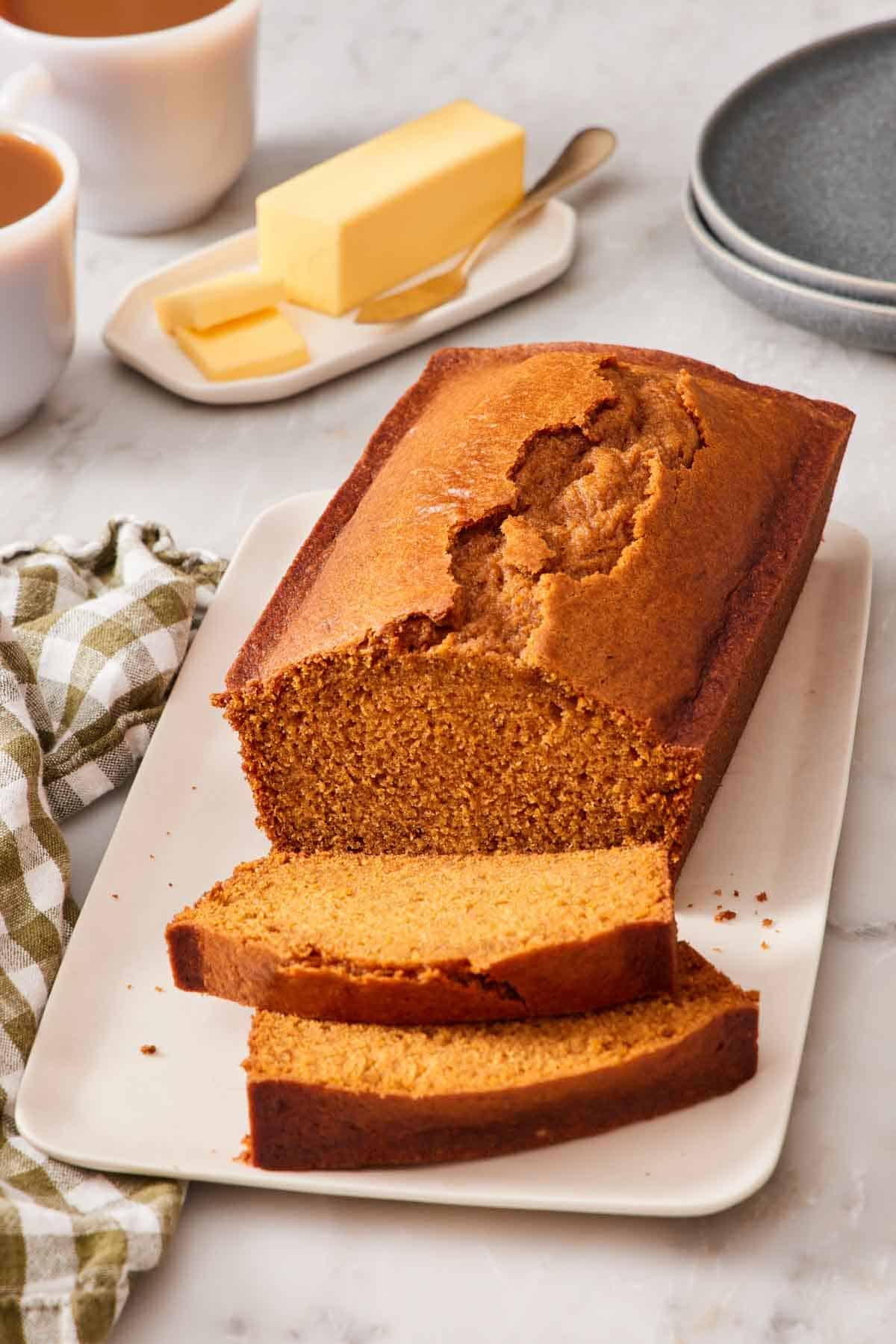 A partially sliced loaf of pumpkin bread on a platter with a stick of butter in the background.