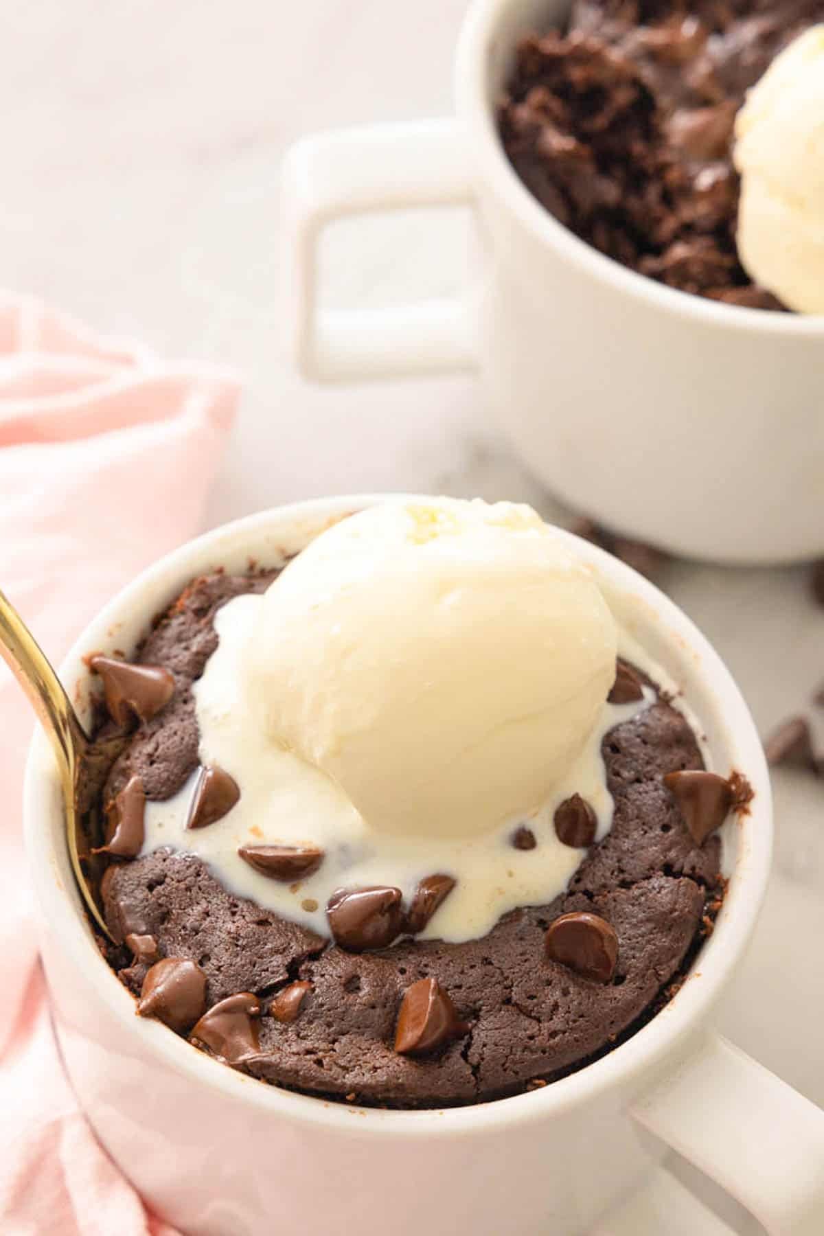 Close-up of a brownie in a mug with a scoop of ice cream on top. Another mug brownie mostly out of focus in the background.