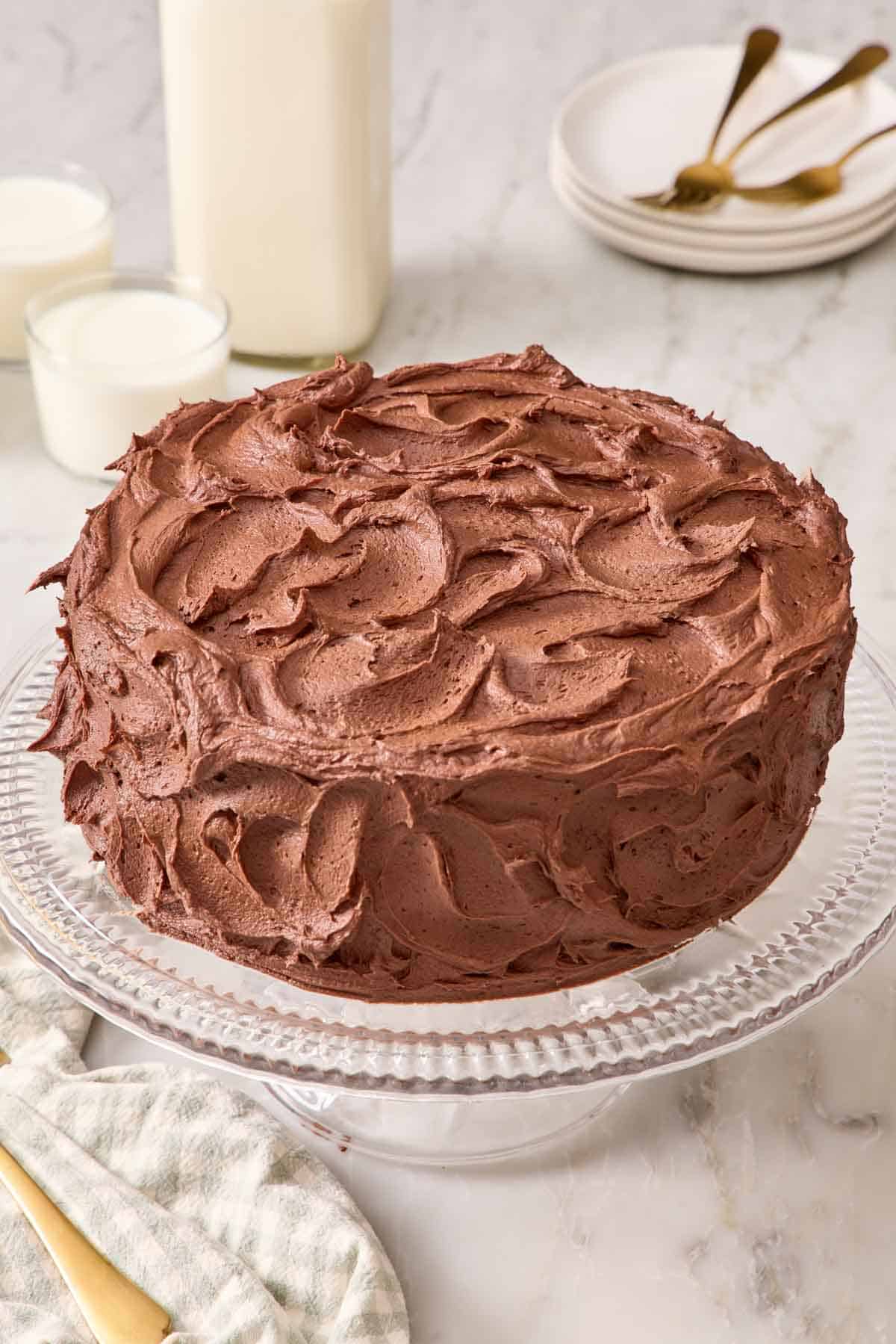 A frosted chocolate cake on a glass cake stand.