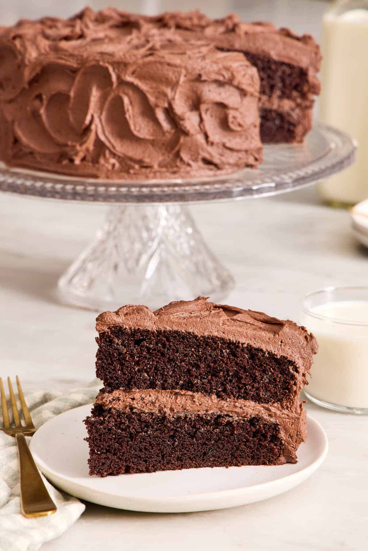 A slice of chocolate cake on a small plate with the rest of the cake on a stand in the background.