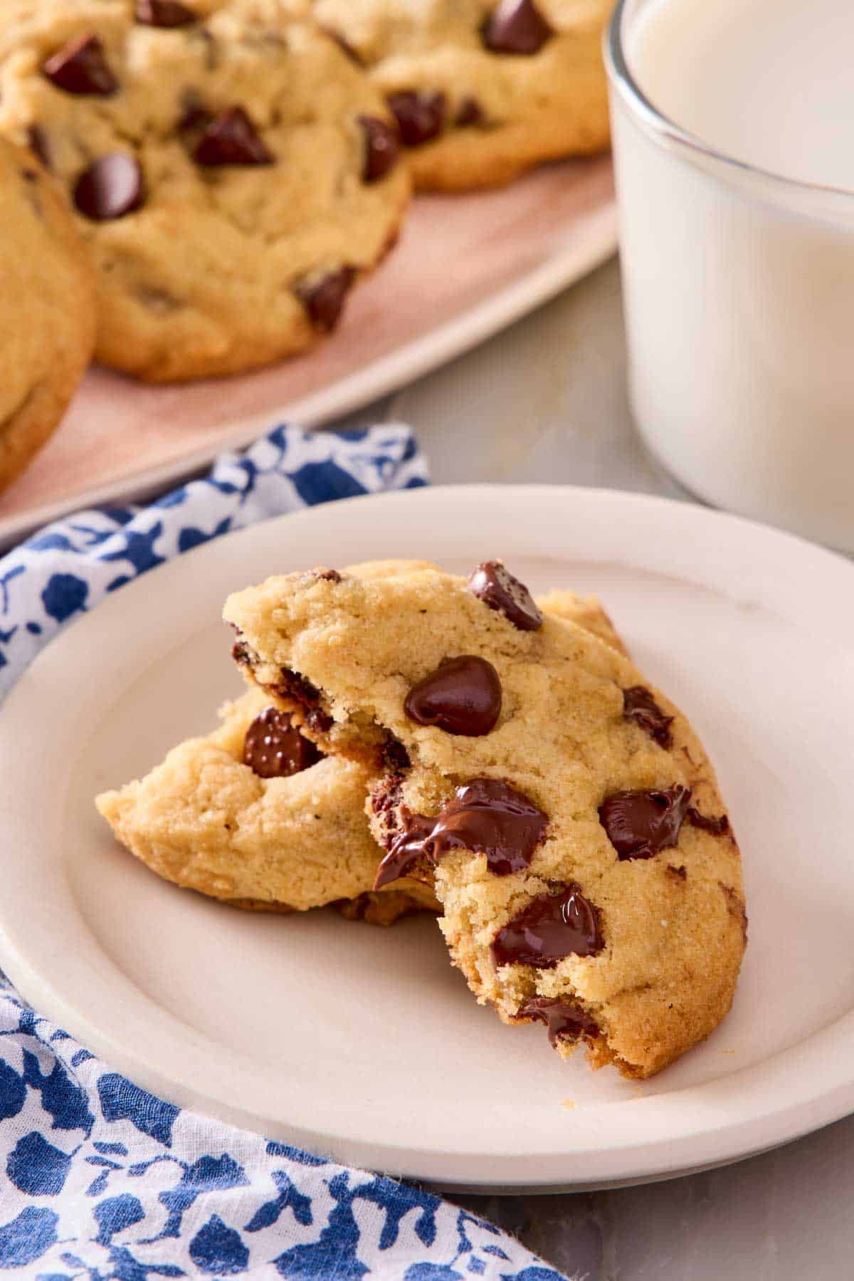 A choc chip cookie torn in half and placed on a small plate with a glass of milk alongside. More cookies in the background.