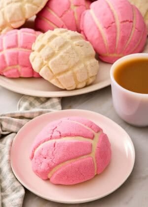 A pink concha on a plate with a cup of coffee and more conchas in the background.