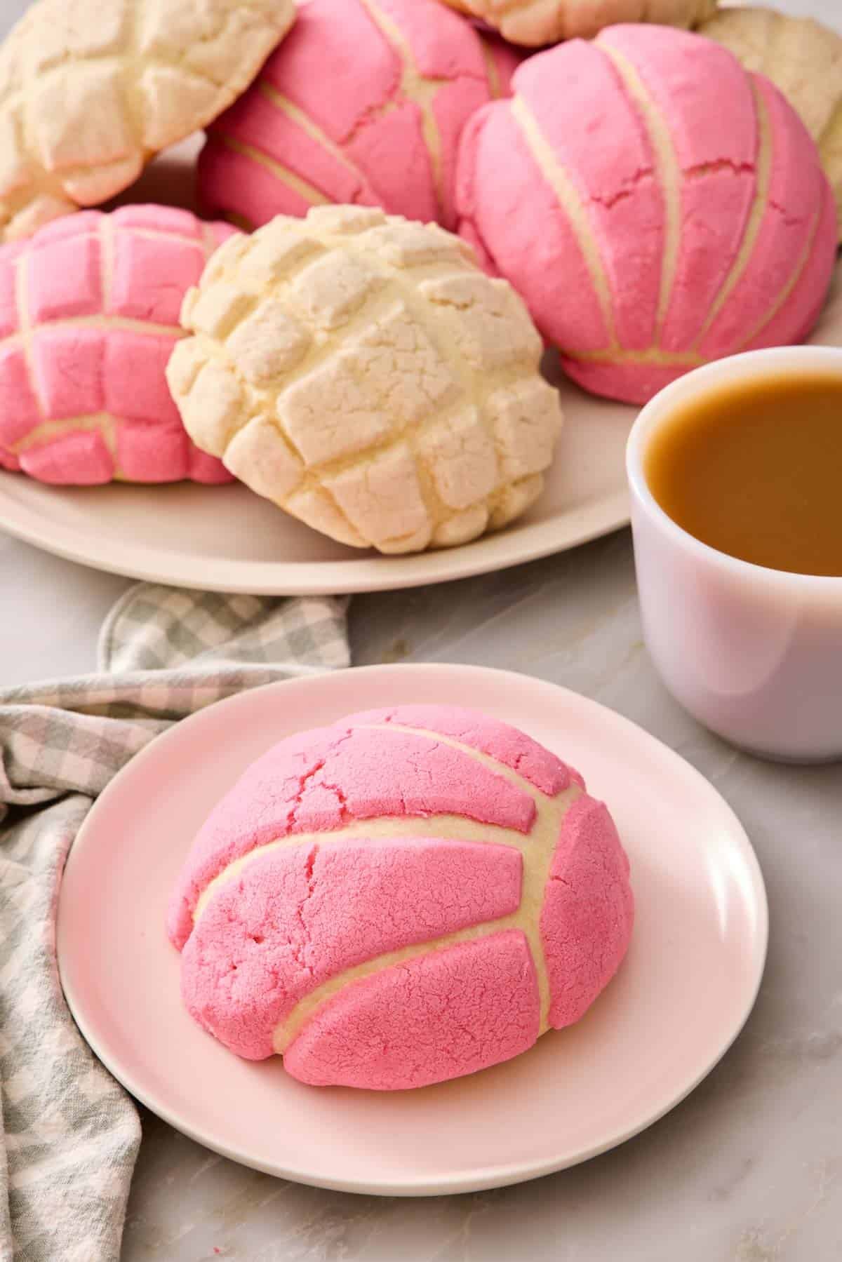 A pink concha on a plate with a cup of coffee and more conchas in the background.