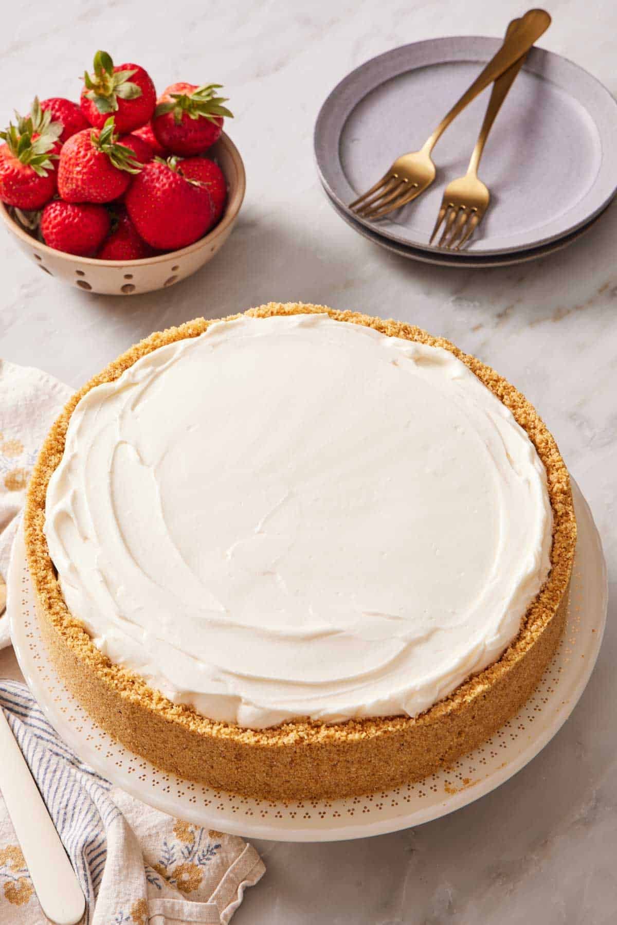 A no bake cheesecake on a stand with a bowl of strawberries and plates behind it.
