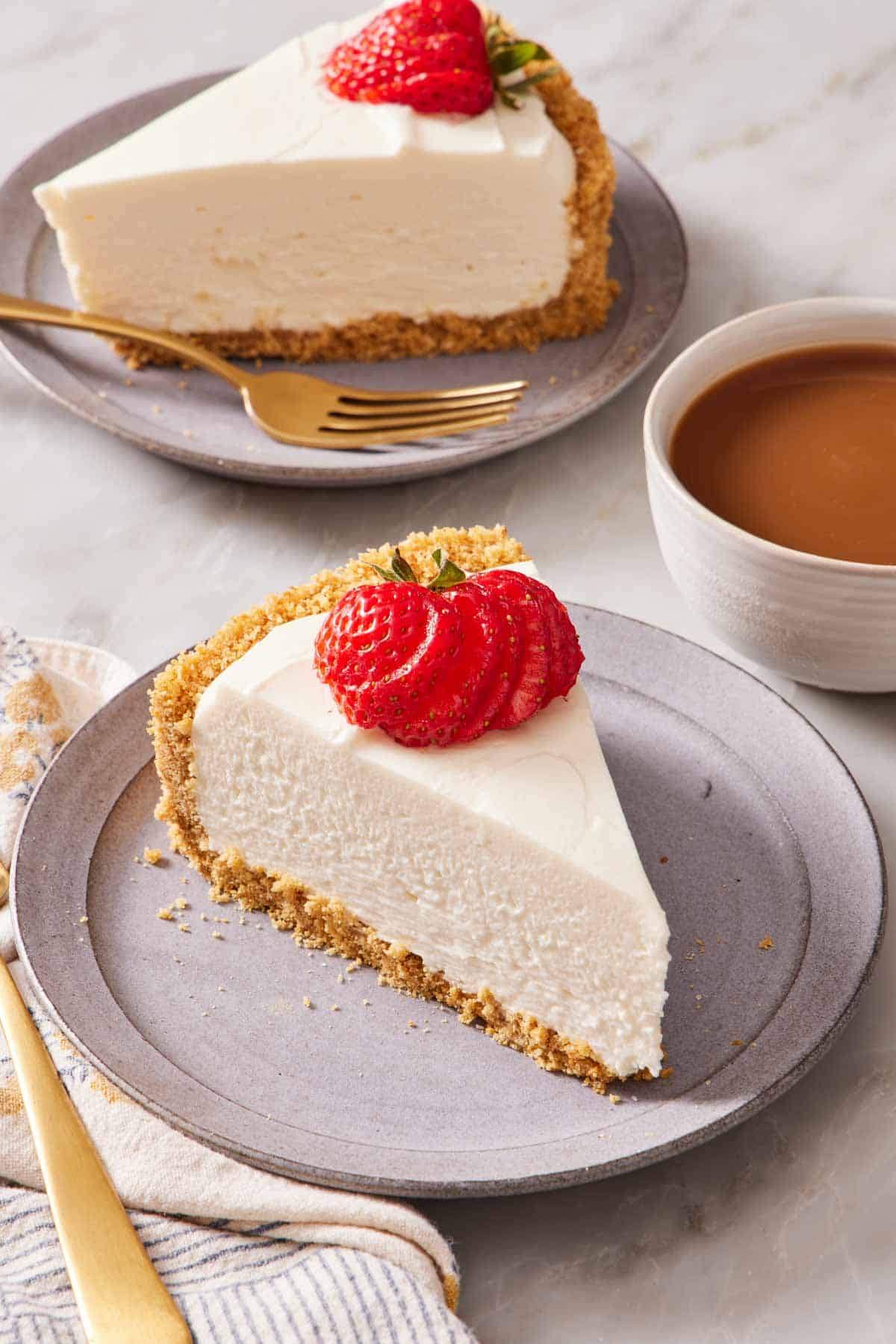 A slice of no bake cheesecake topped with a strawberry next to a cup of coffee. Another slice in the background.