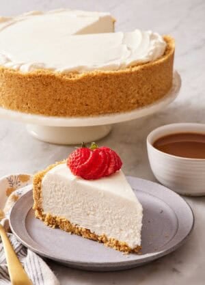 A slice of no-bake cheesecake on a plate next to a cup of coffee. The rest of the cheesecake on a cake stand behind it.