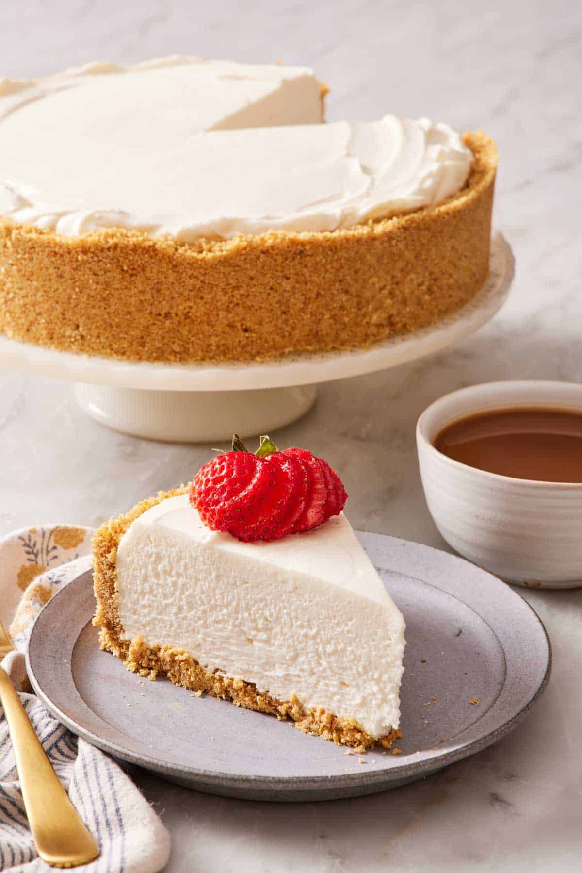 A slice of no-bake cheesecake on a plate next to a cup of coffee. The rest of the cheesecake on a cake stand behind it.