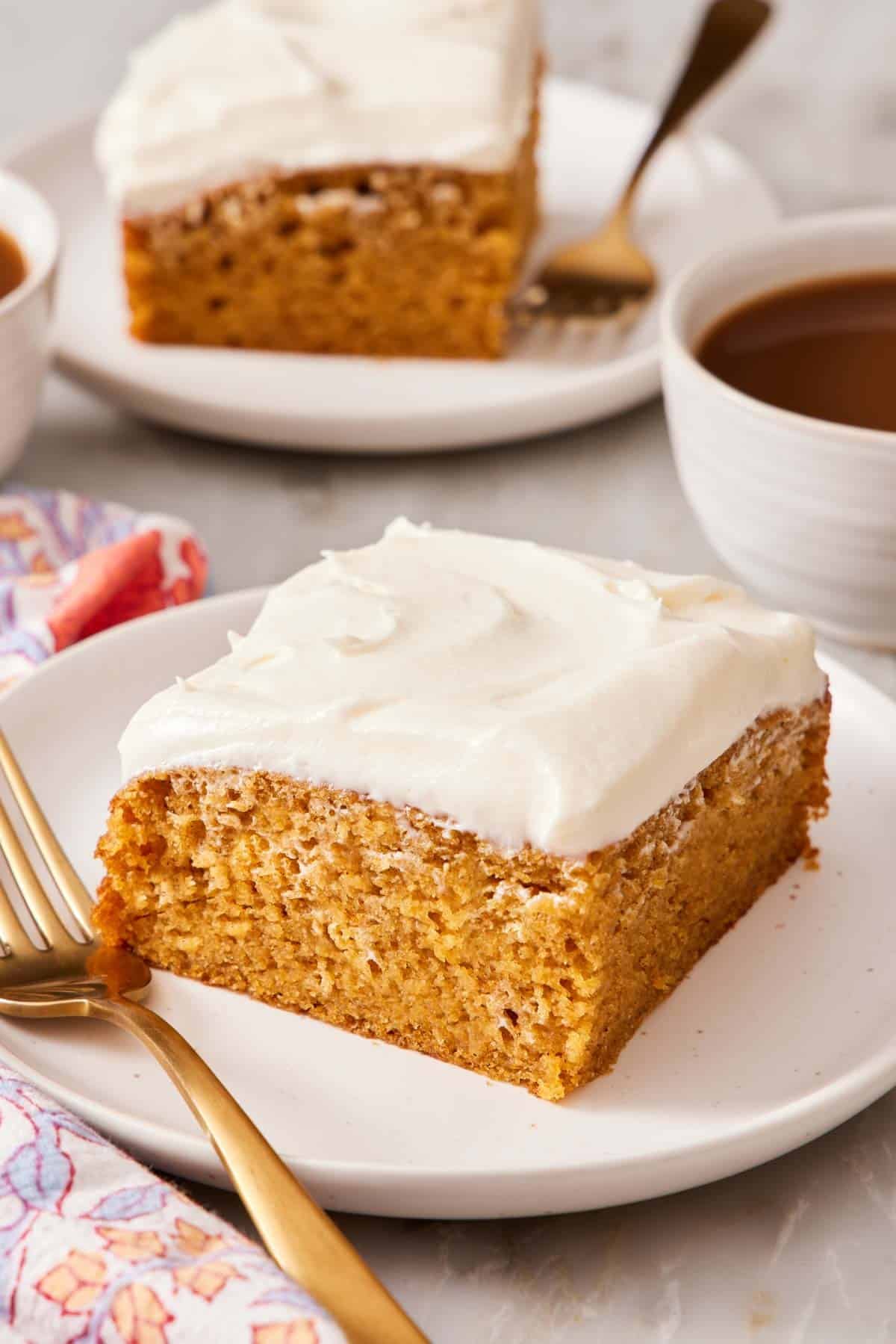 A slice of pumpkin cake on a small plate with a work and a cup of coffee and more cake behind it.