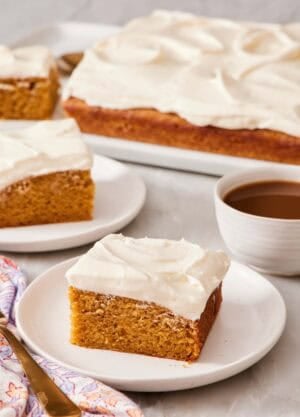 Slice of pumpkin cake on a plate alongside a cup of coffee and more slices of cake, as well as the rest of the cake.