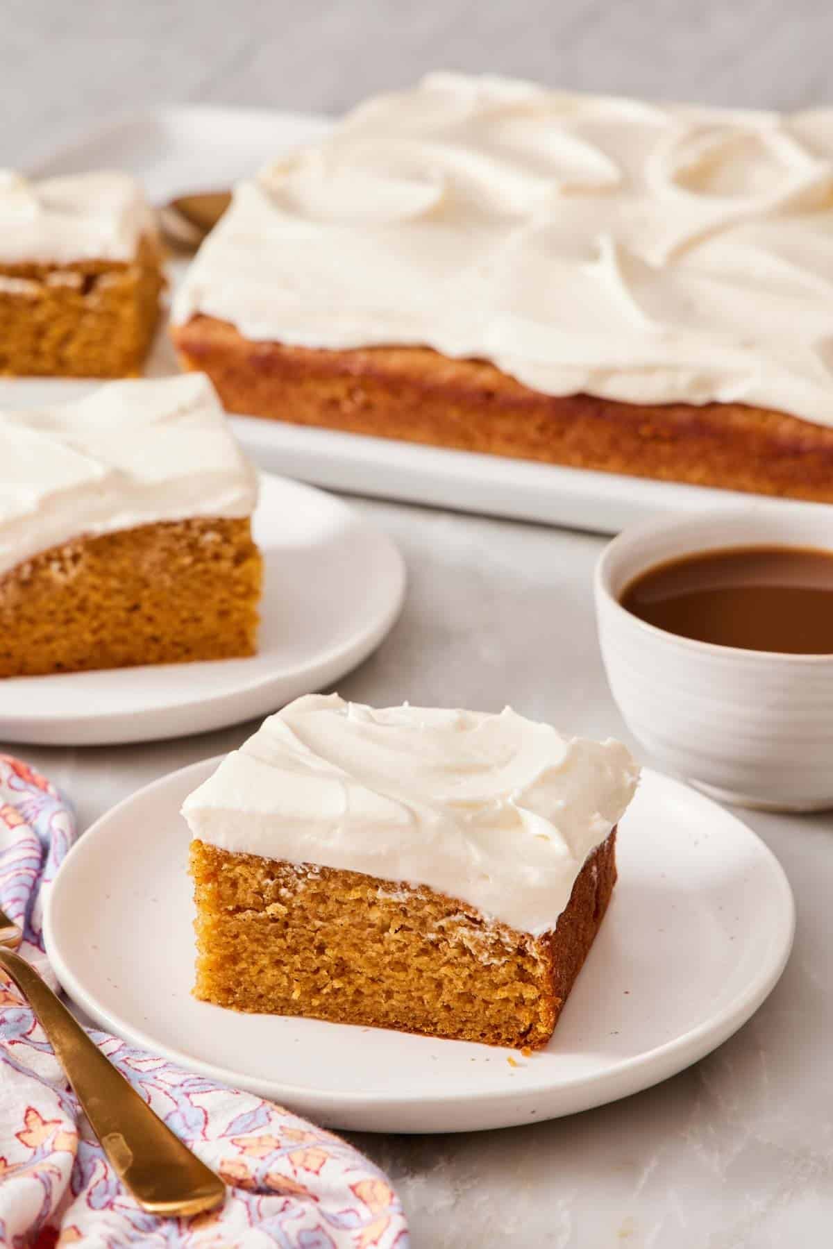 Slice of pumpkin cake on a plate alongside a cup of coffee and more slices of cake, as well as the rest of the cake.