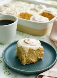 A plate with a cinnamon roll. A baking dish with more rolls and a mug of coffee in the back.