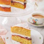 A plate with a slice of Victoria sponge cake in front of a cup of coffee and the rest of the cake.