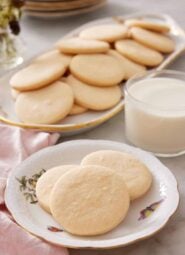 A plate with three sugar cookies with a glass of milk and additional cookies on a platter in the background.