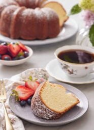 A plate with a slice of cream cheese pound cake along with some berries, dusted with powdered sugar. A drink, more berries, and the rest of the cake in the background.