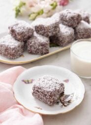 A plate with a lamington with a glass of milk and platter of more lamingtons.