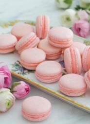 A group of pink macarons on a serving tray next to flowers