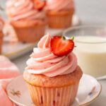 A strawberry cupcake with half a strawberry on top of the pink frosting. More cupcakes and milk in the background.