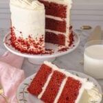 A cake stand with a slice of red velvet cake cut out and placed on a plate in front.