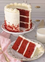 A cake stand with a slice of red velvet cake cut out and placed on a plate in front.